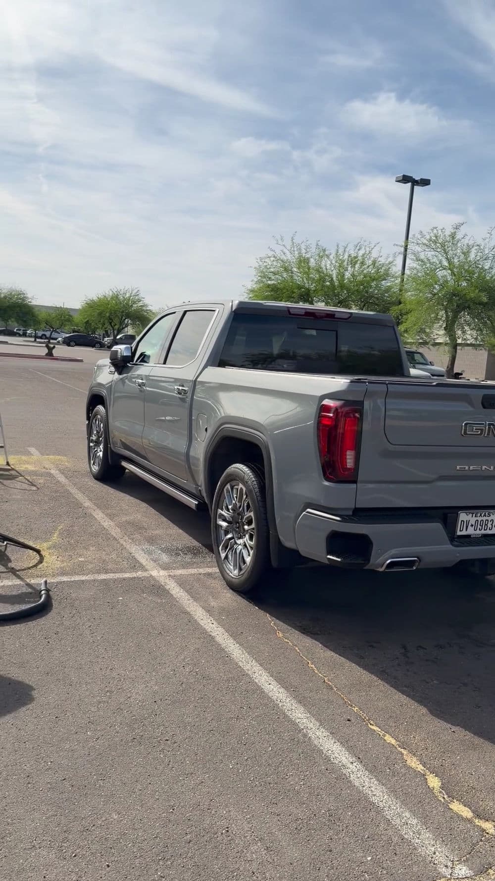 Gray GMC truck parked at a charging station in an outdoor lot with trees in the background.
