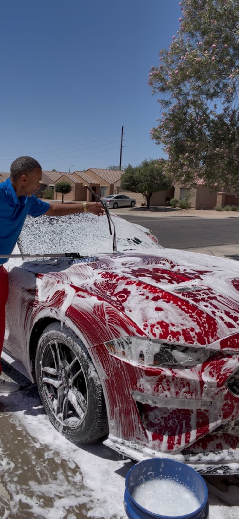 Man washing a red car with foam in a residential area on a sunny day.