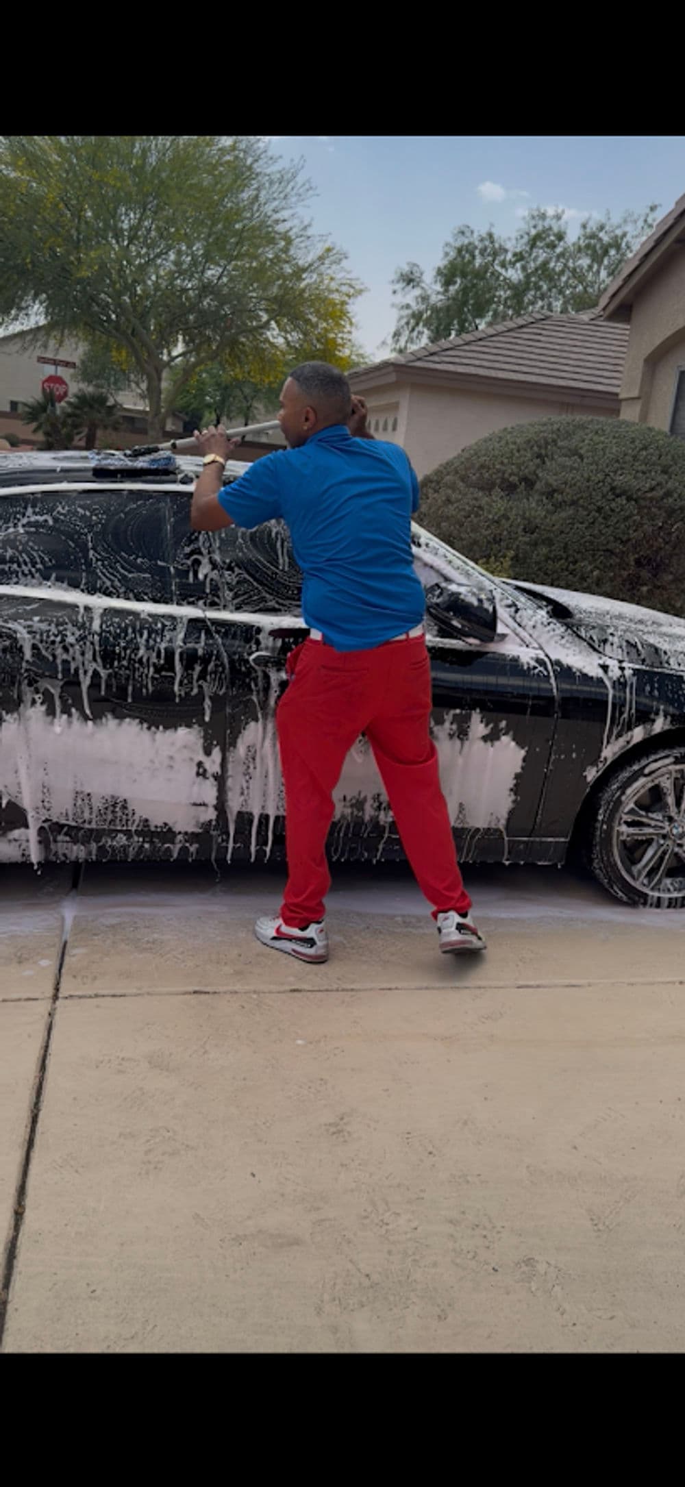 Person washing a black car covered in soap outdoors, wearing a blue shirt and red pants.