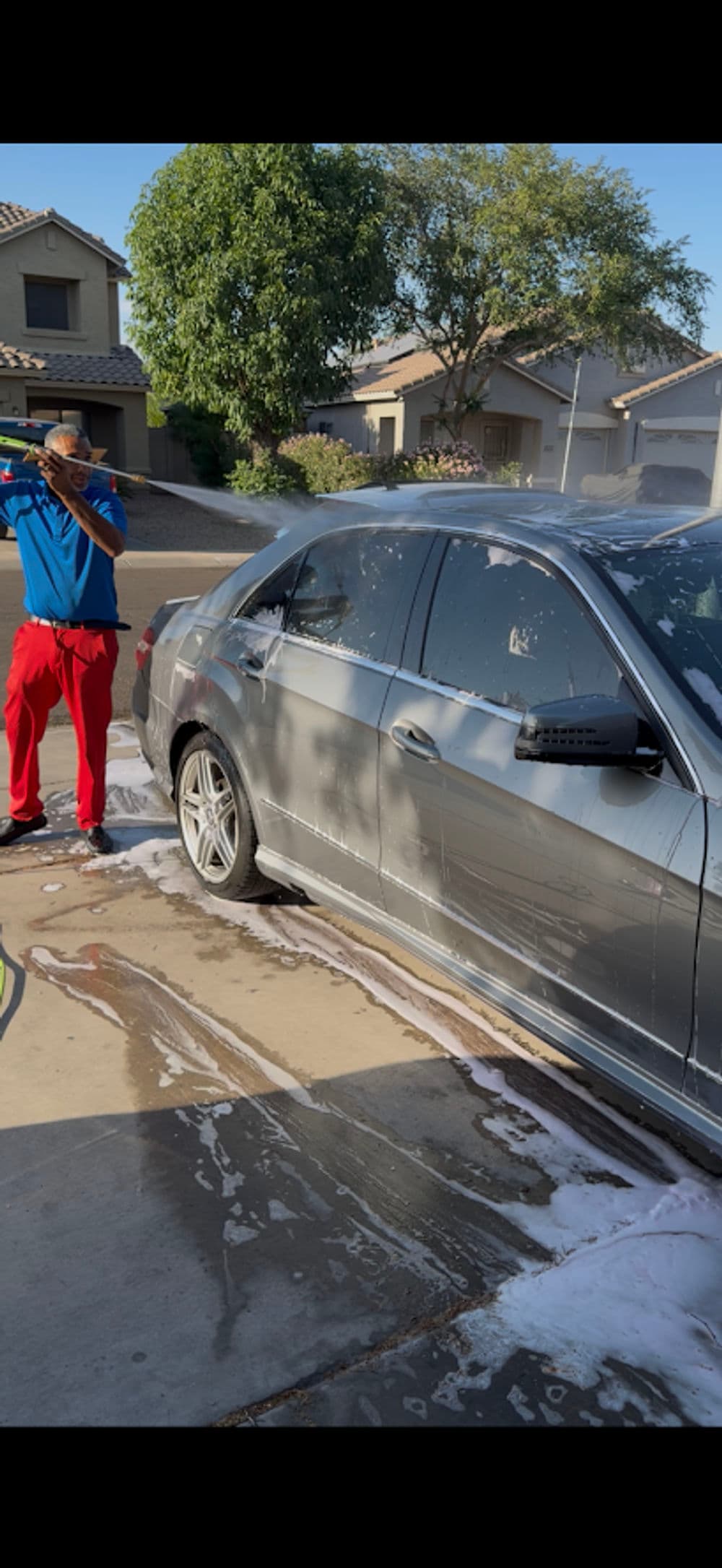 Man washing a gray car with soap in a residential driveway on a sunny day.